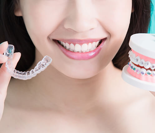 woman holding a clear aligner up next to a model of teeth with traditional braces