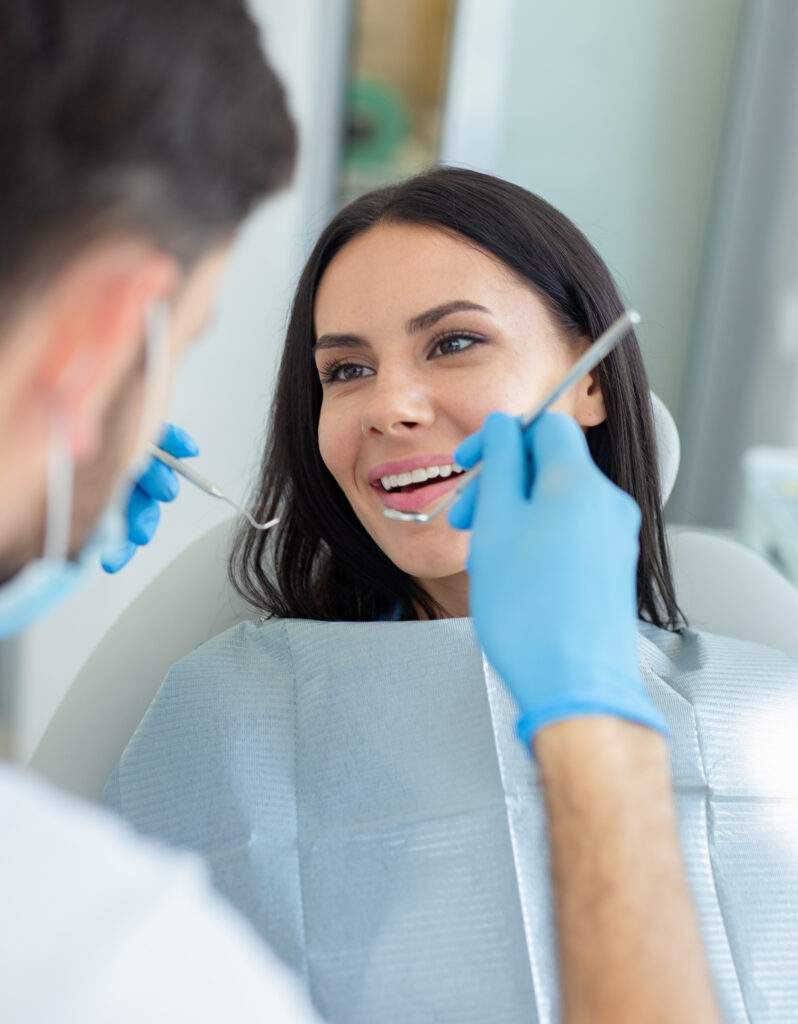 A dentist wearing gloves examines a smiling woman’s teeth with dental tools while she sits in a dental chair.