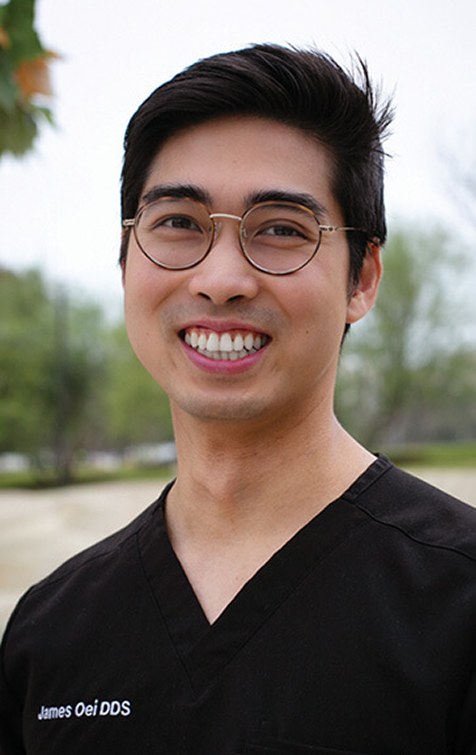 A man with glasses and dark hair, wearing black scrubs with the text "James Oei DDS" on them, smiles outdoors with trees in the background.